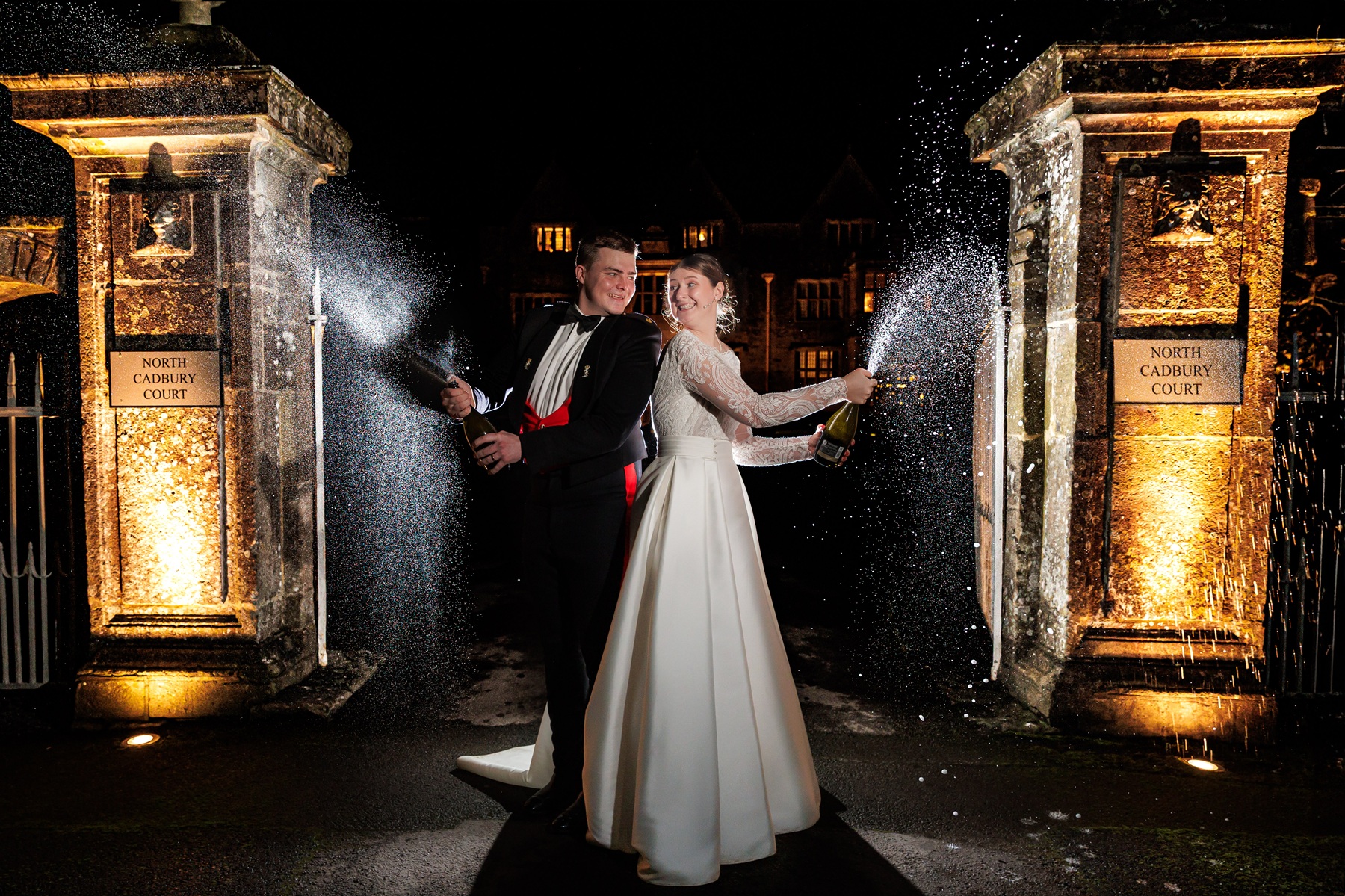 bride and groom standing back to back in a lit up ornamental drive way with champagne spraying while looking at each other and laughing
