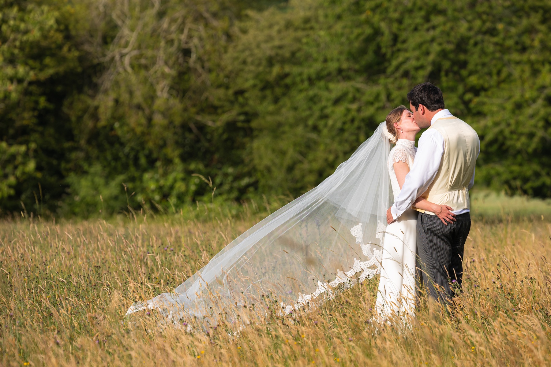 bride and groom in long grass, facing each other, arms around each other, kissing
