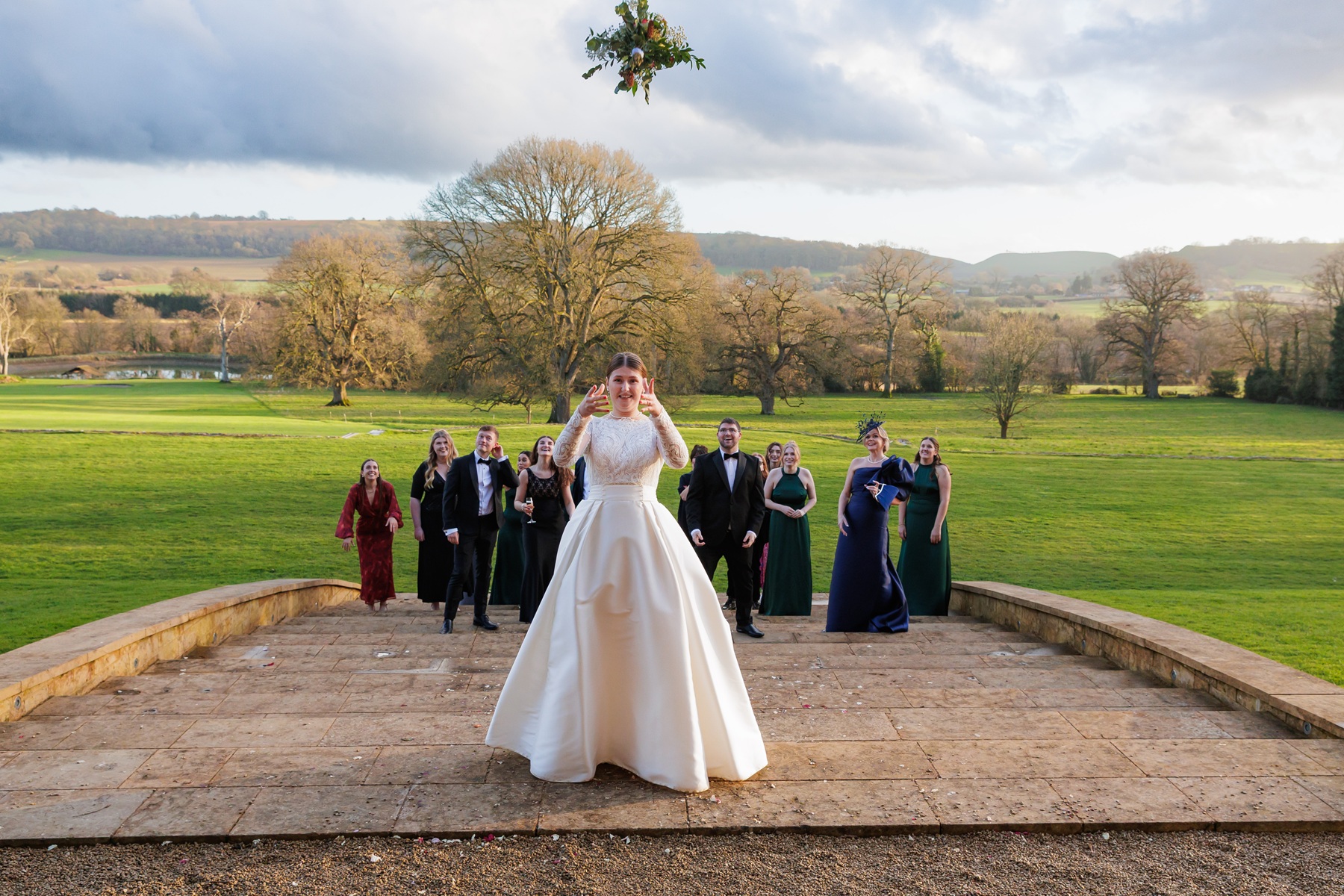 bride standing at the top of outdoor staircase throwing her bouquet backwards to waiting friends