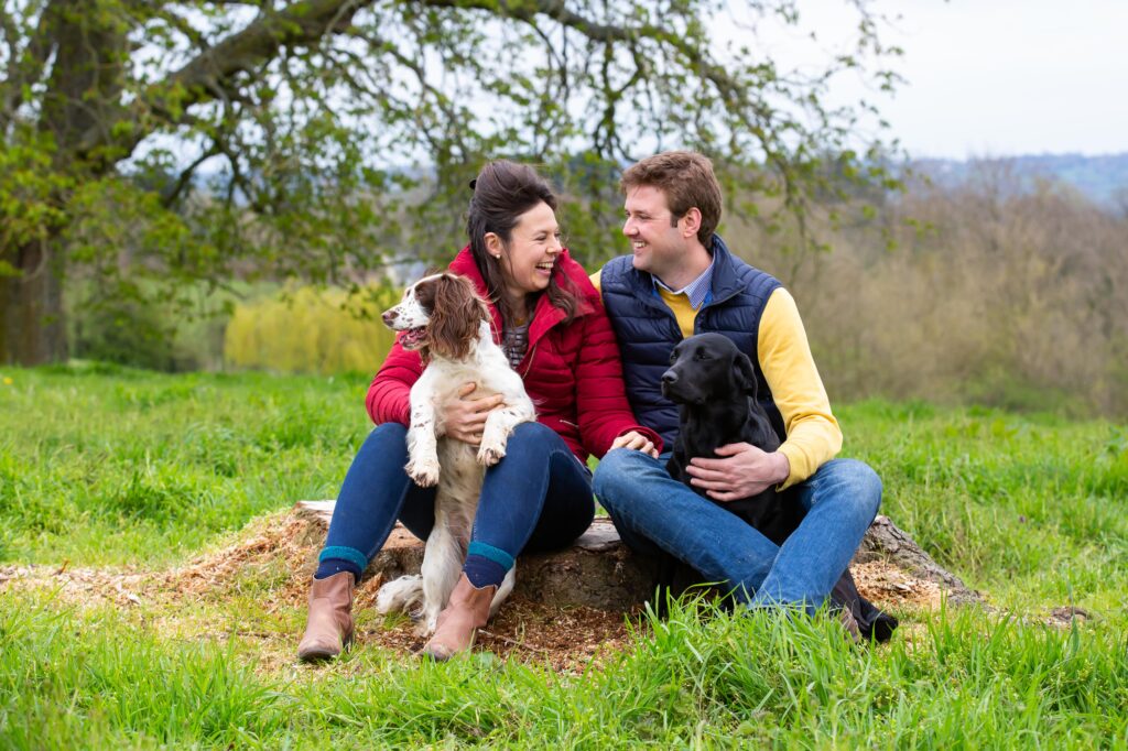 engaged couple sitting on a tree stump facing each other laughing with their dogs