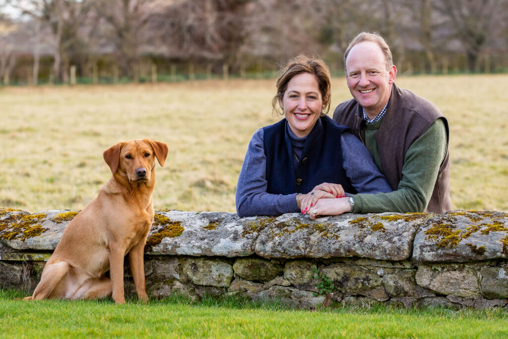 engaged couple leaning on a wall clasping hands with their dog sitting close