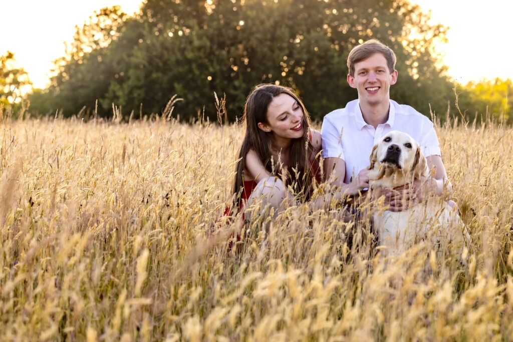 engaged couple sitting in long grass with their dog during golden hour