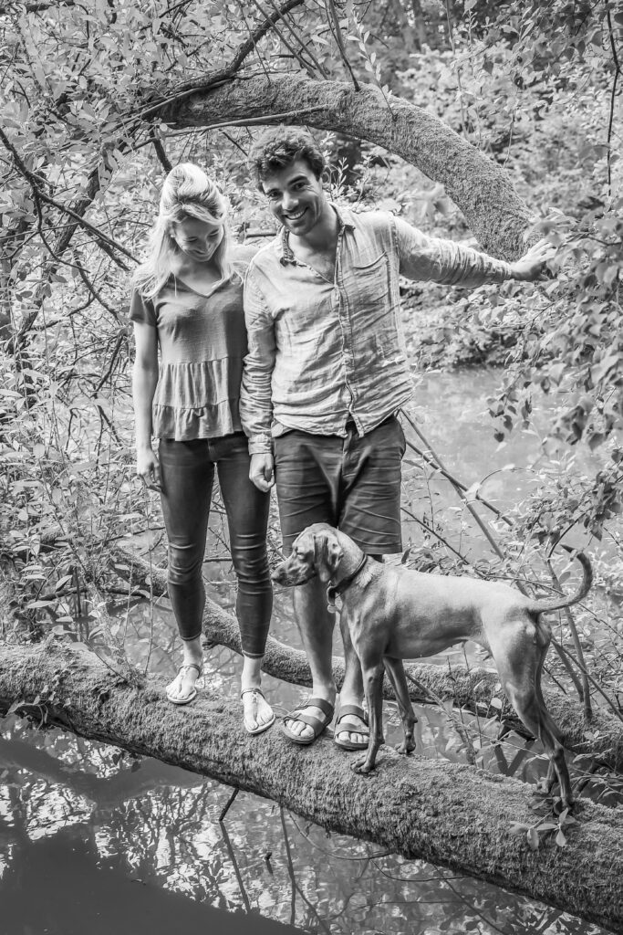 engaged couple standing on a fallen tree over a pond with their dog